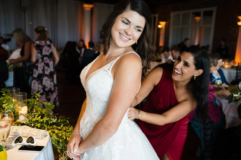 Laurel in her white beaded wedding dress laughs as a guest in burgundy adjusts the back of her gown during the reception.