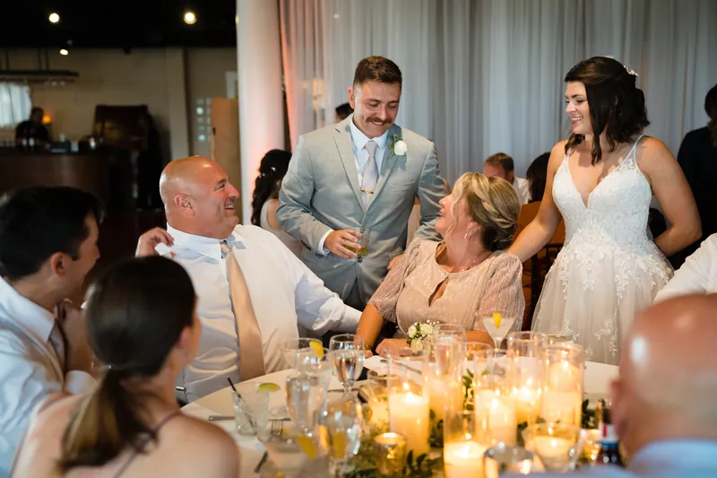 Jake holds a microphone while standing at a dining table, speaking to Laurel and assembled guests during the reception.