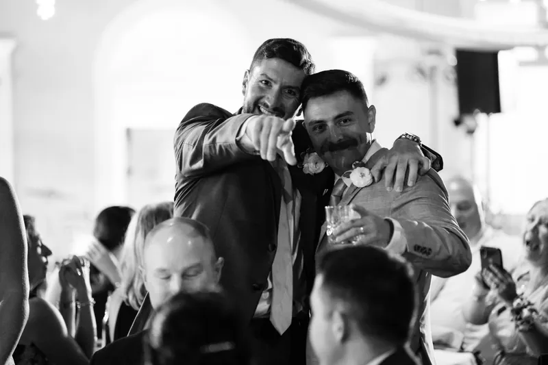 Two men embrace warmly on a crowded dance floor while guests surround them in a black-and-white photograph.