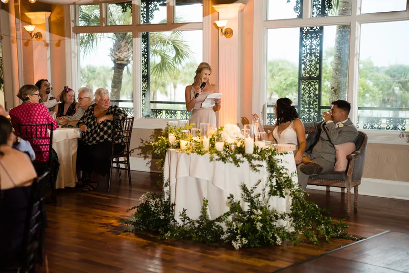 Laurel speaks from a microphone at a draped table adorned with greenery and candles, facing seated guests near tall windows.