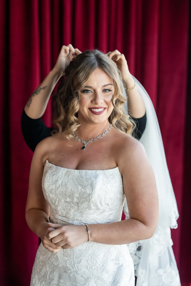 Lauren holds her veil at the back of her head, smiling in a strapless white lace gown against a deep burgundy curtain.