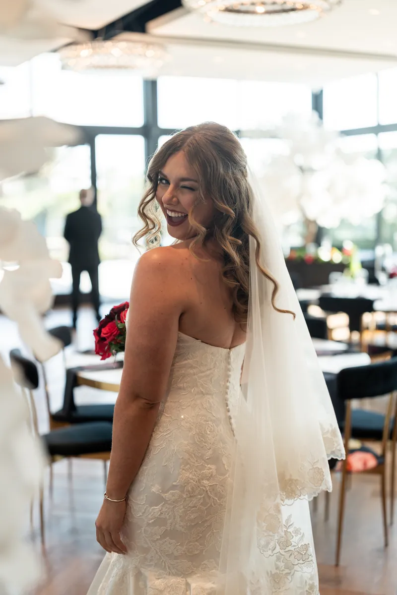 Lauren turns toward the camera in her ivory lace wedding dress with a veil trailing behind her in the reception space.