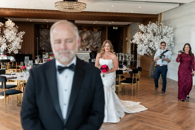 Kevin stands in a black tuxedo with a bow tie as Lauren walks past him in her white gown across the wood dance floor.