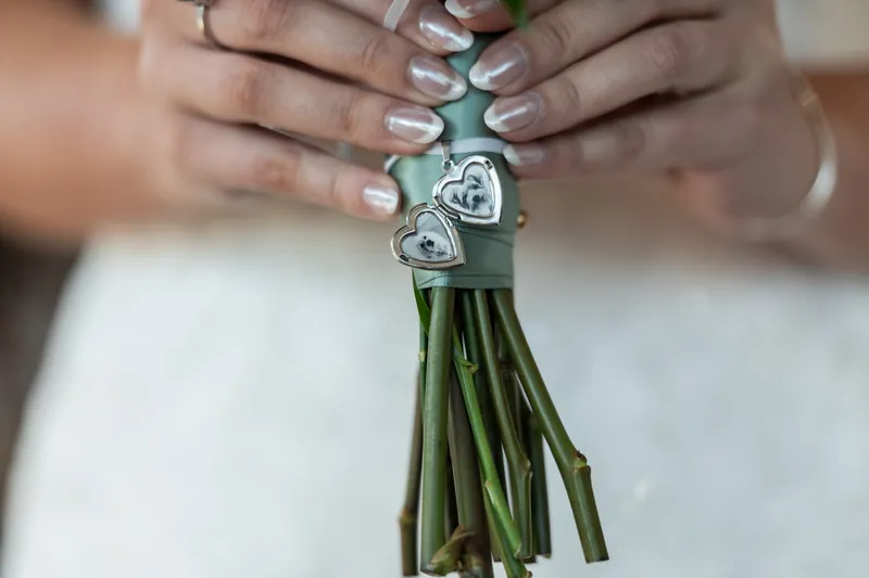 Hands display a bridal bouquet wrapped with decorative silver charms shaped like hearts against a white dress.