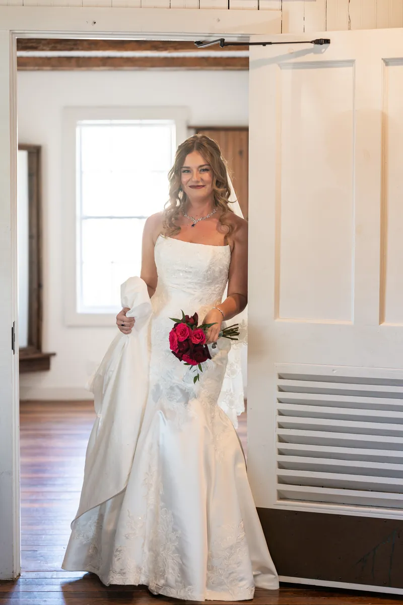 Lauren poses alone in a chapel hallway in her strapless white gown, holding her crimson bouquet near a bright doorway.