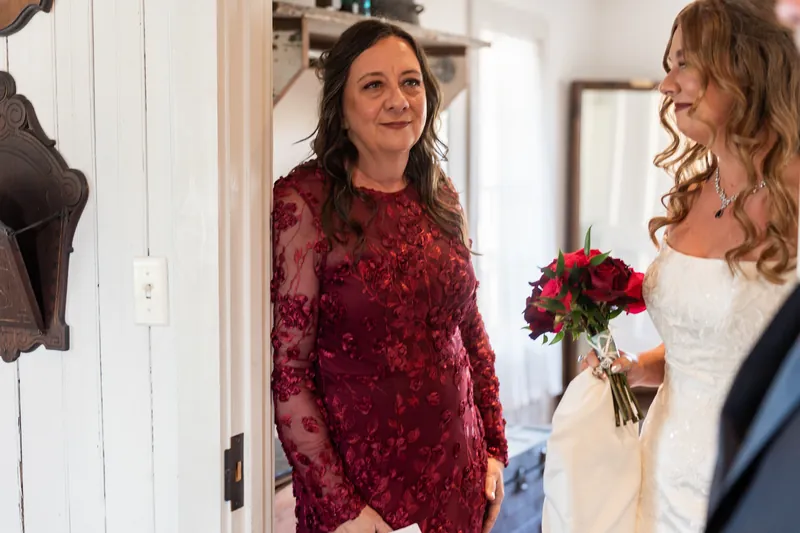 A woman in burgundy beams at Lauren, who holds her red bouquet in the chapel's white interior.