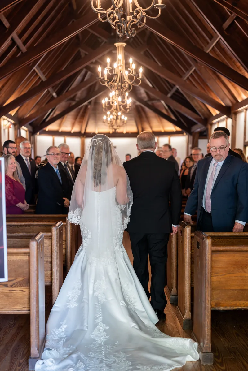 A bride in a flowing white gown walks the aisle toward the altar as guests observe.