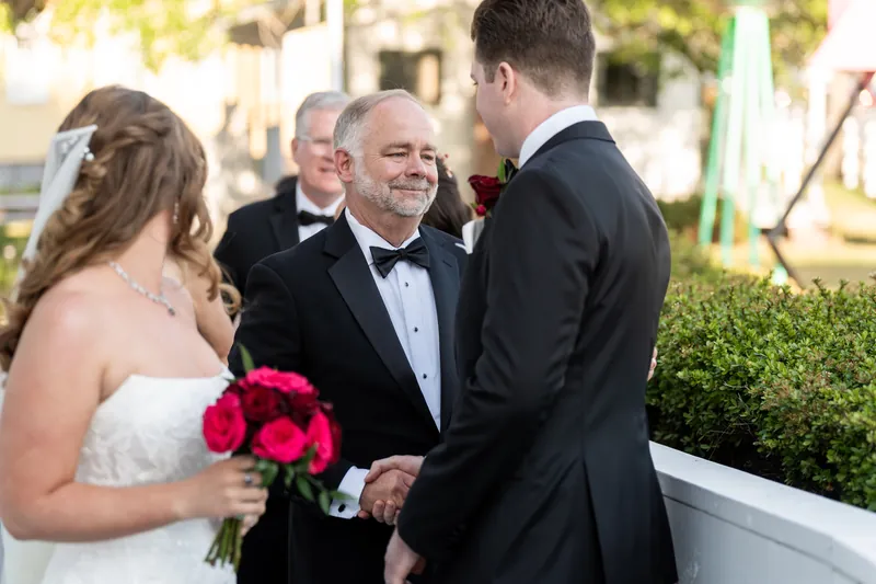 Kevin holds Lauren's hand while speaking with the officiant outside the chapel, Lauren clutching her hot pink bouquet.
