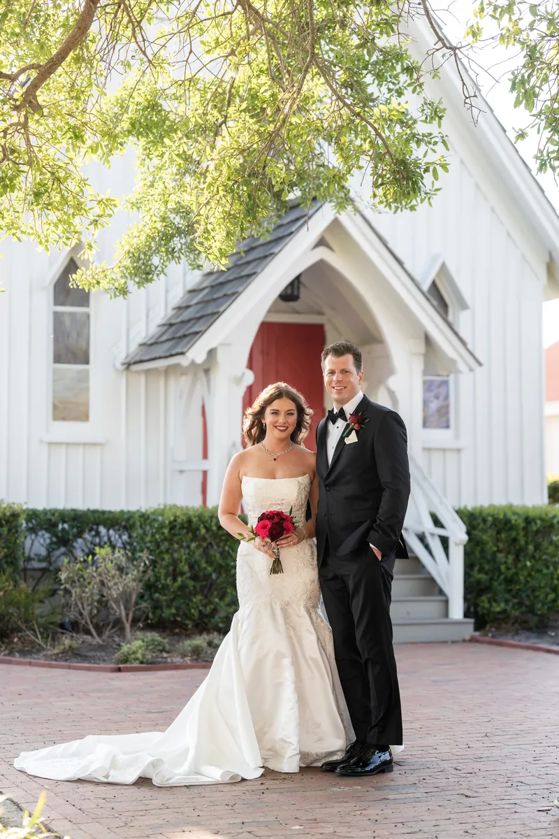 Lauren and Kevin pose in front of the white chapel with its red doors, Lauren carrying her bouquet and trailing veil.