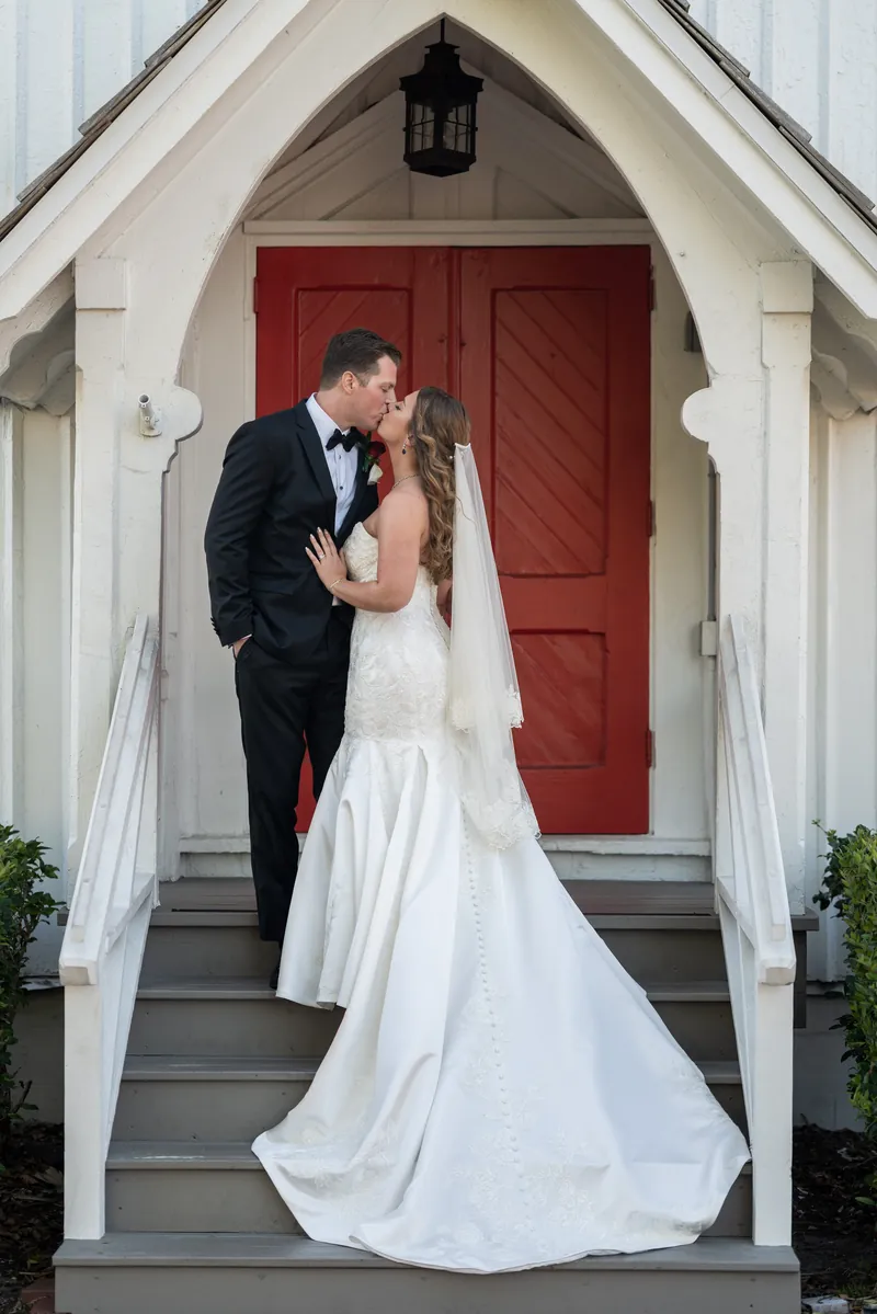 Kevin and Lauren kiss on the chapel steps beneath the red entrance door, Lauren's white gown and veil flowing.