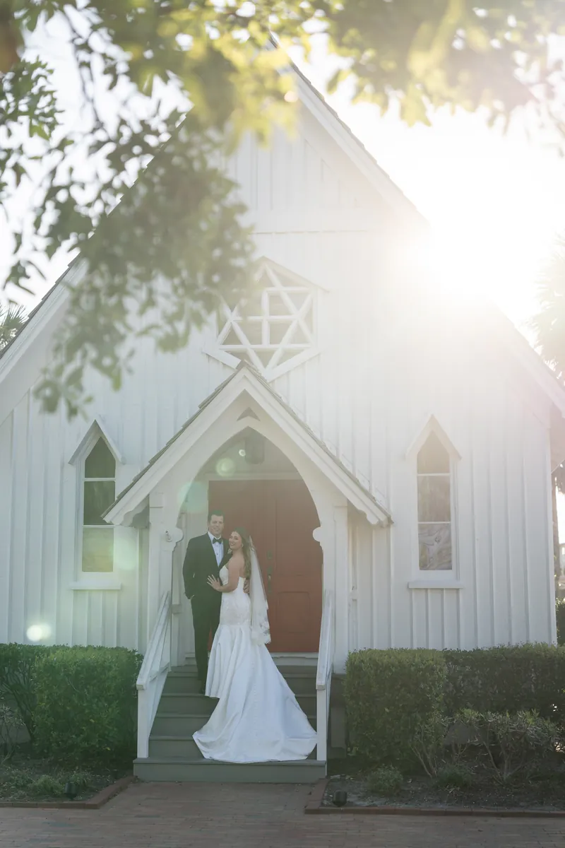 Lauren and Kevin stand in the chapel doorway at golden hour, sunlight filtering through trees overhead.