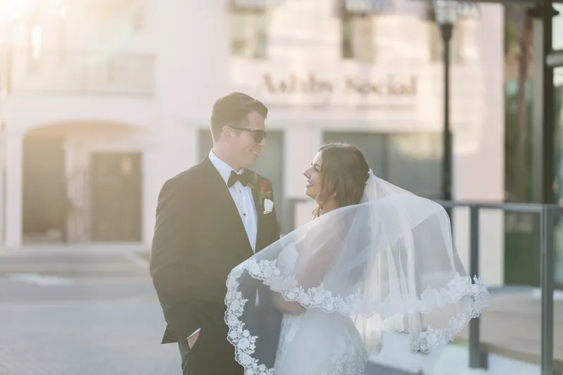 Kevin in a dark suit with a boutonniere gazes at Lauren, who wears a white lace gown and veil, standing together outdoors.