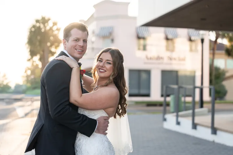 Kevin wraps his arms around Lauren from behind while she laughs, both dressed formally with modern buildings and landscaping
