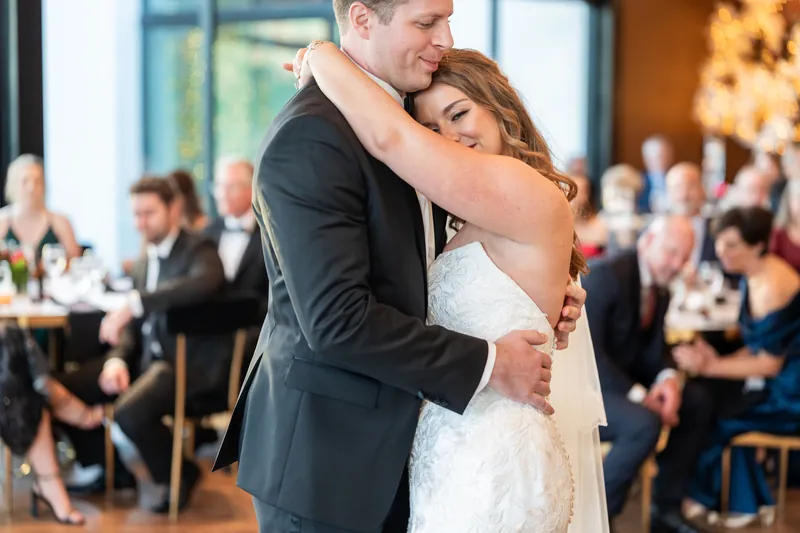 Lauren rests her head on Kevin's shoulder while they slow dance, surrounded by seated guests in the softly lit reception