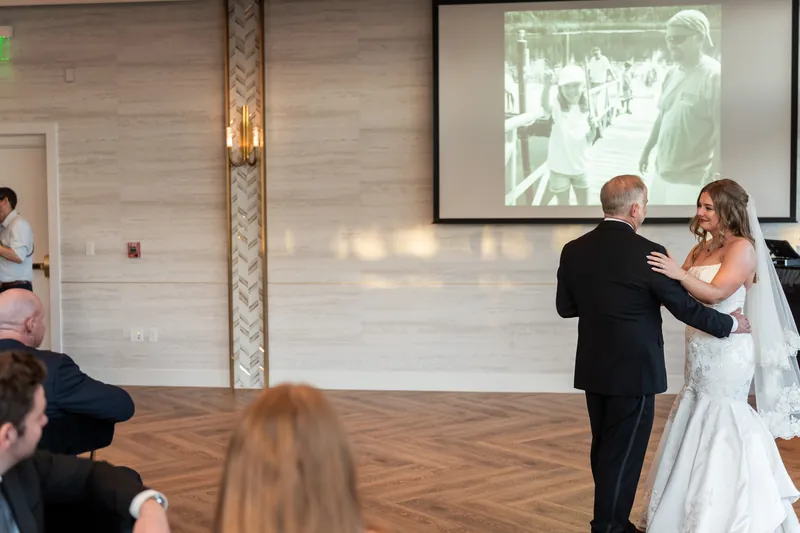 Kevin and Lauren dance together as a video plays on a screen behind them and guests observe from the wooden dance floor.