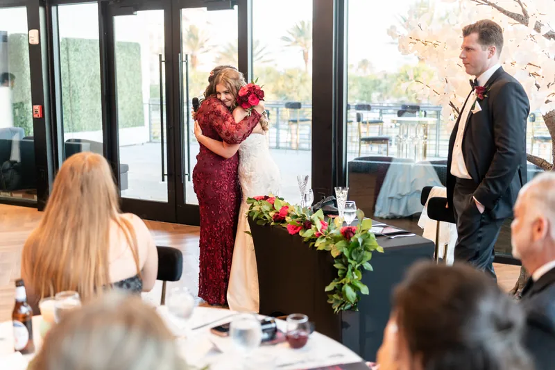 A woman in a burgundy gown speaks at the microphone while Kevin stands nearby, with floor-to-ceiling windows and outdoor