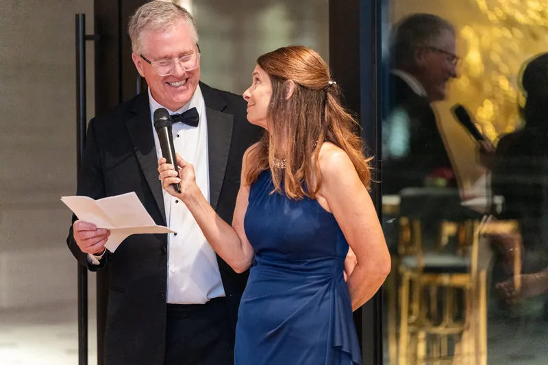 A man reads from a card while speaking with a woman in a blue dress during the evening's toasts.