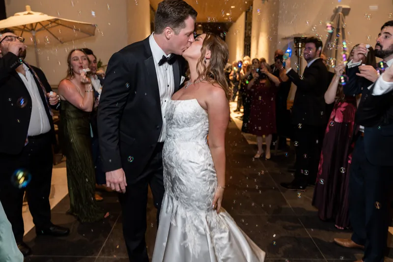 Kevin and Lauren embrace and kiss while surrounded by smiling wedding guests in formal wear.