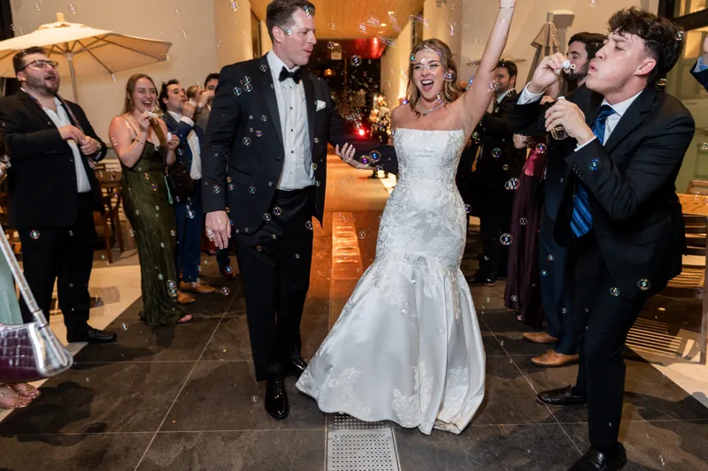 Kevin and Lauren walk across the dance floor while a guest holds a microphone, surrounded by formally dressed wedding