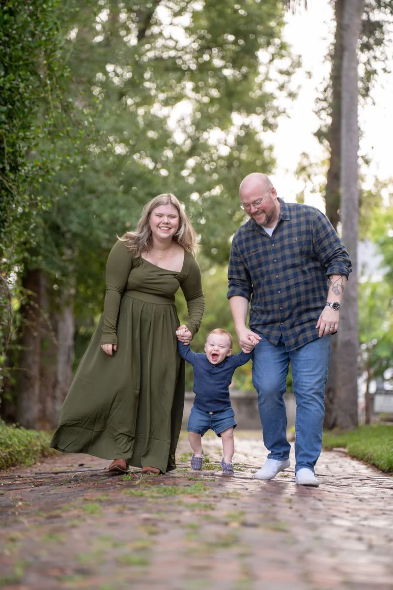 Justin and Lauren stand together on a brick path, both smiling while holding their son Casen between them.