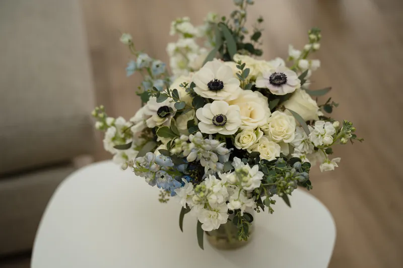 White anemones, garden roses, and greenery arranged in a clear glass vase on a white table.