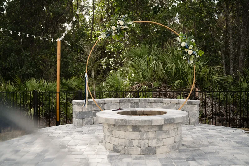 Circular stone fire pit surrounded by pavers, framed by a metal arch draped with white florals and greenery.