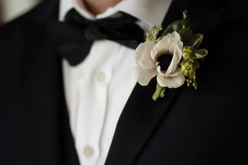 Person in formal black tuxedo with white shirt and bow tie wearing a white anemone boutonniere.