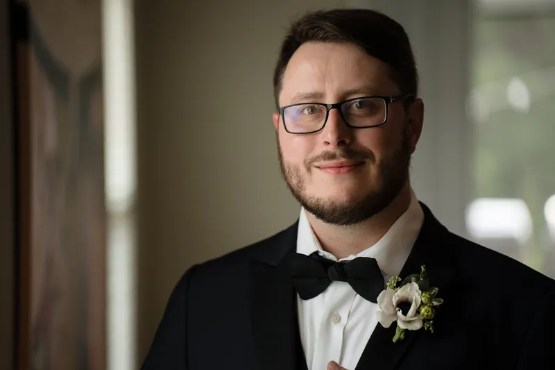 Marco smiles at the camera in a black tuxedo, white dress shirt, and bow tie with a matching boutonniere.