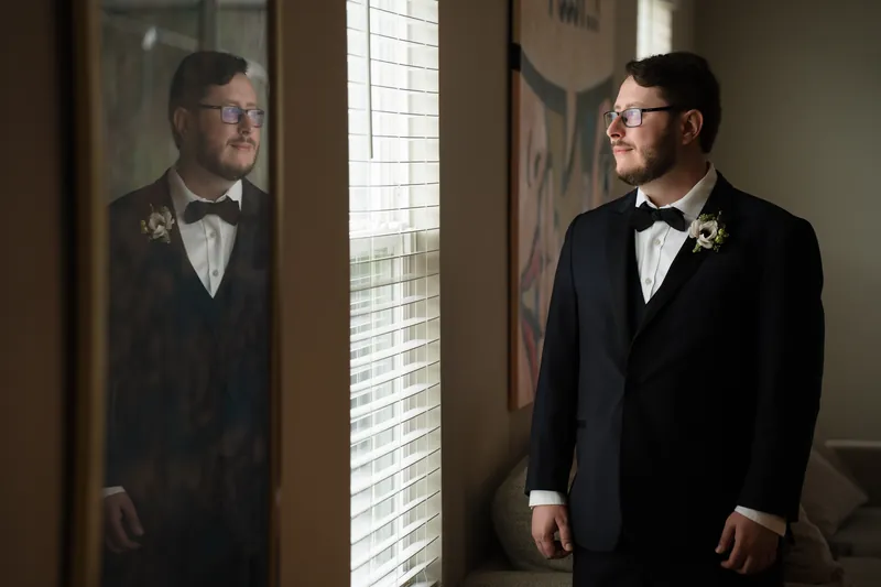 Two people in black tuxedos and bow ties stand facing each other near a window with gridded panes.