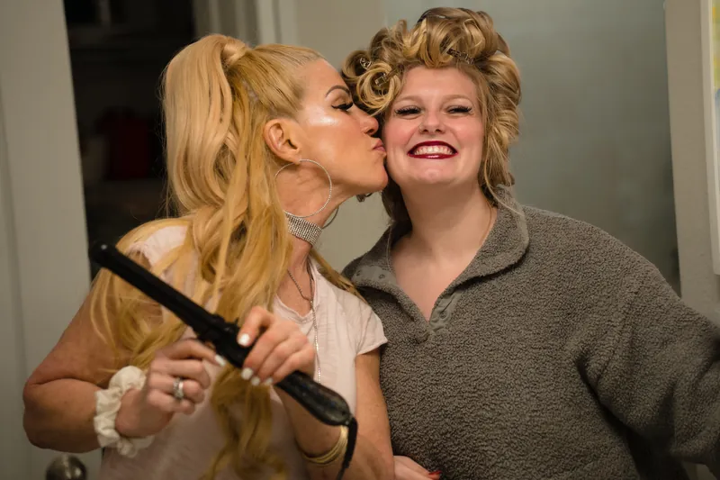One person in a cream top kisses another person's cheek while holding a makeup brush during wedding preparations.