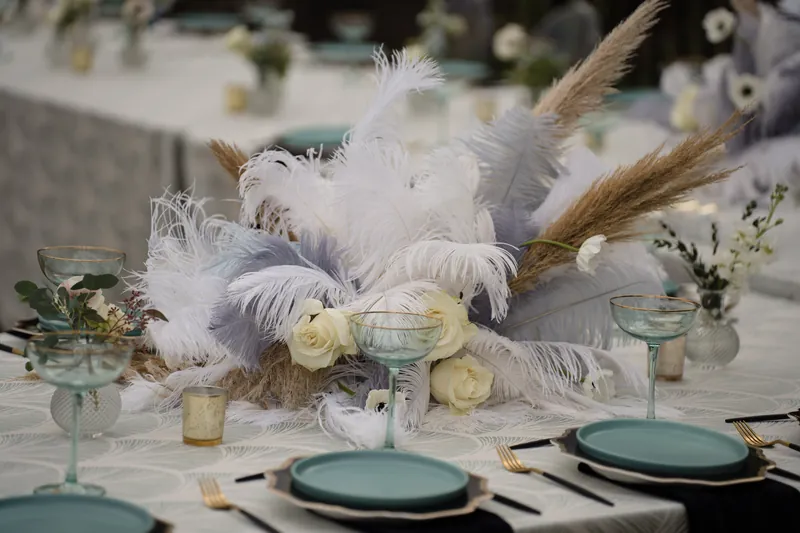 Elegant table setting features cream roses, white feathered arrangements, teal plates, and greenery in glass vases.