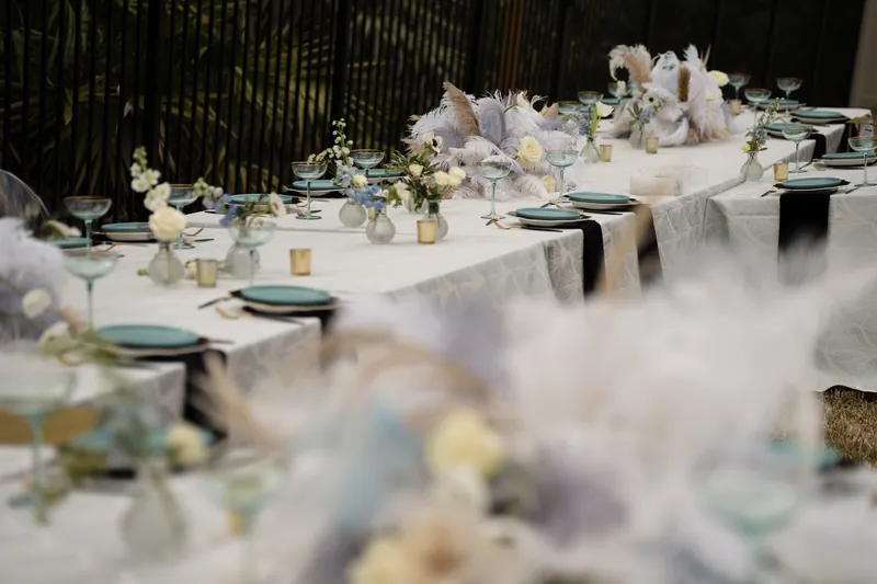 Long white reception table set with pale florals, black napkins, and glassware under warm lighting.