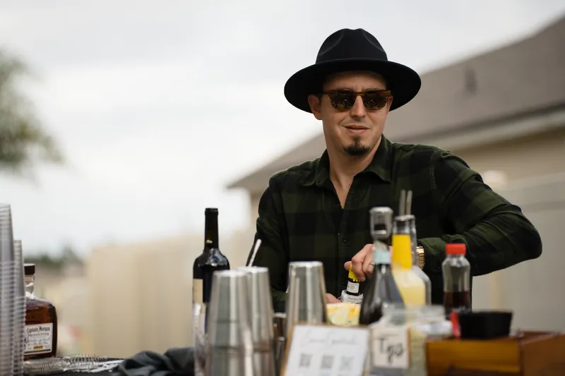 Person in dark shirt and black fedora stands behind a bar setup with bottles and glassware.