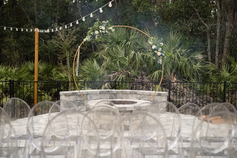 Wooden arch with string lights frames a circular stone fire pit surrounded by palmetto plants and transparent chairs.