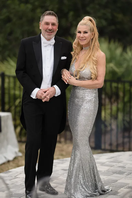 Lea & Marco stand together outdoors, he in a black suit and white shirt, she in a sequined silver gown.