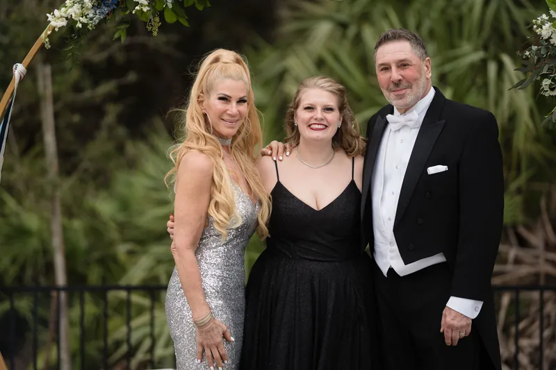 Three people pose under a floral arch with palm fronds, wearing formal attire in neutral tones.
