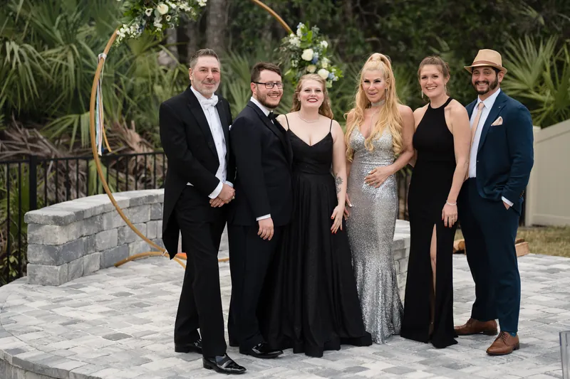 Six guests in formal wear stand on a stone patio with tropical greenery and a wooden arch behind them.