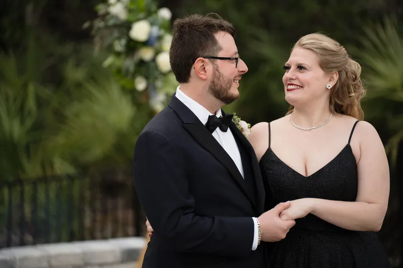 Two people in formal attire smile at each other against a lush green garden backdrop with white flowers.