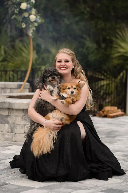 Someone in a black gown kneels on pavement, cradling two small dogs with warm affection.