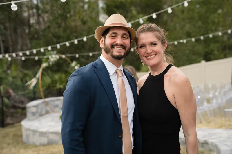 The couple stands under string lights wearing formal attire, with one person in a tan fedora hat.