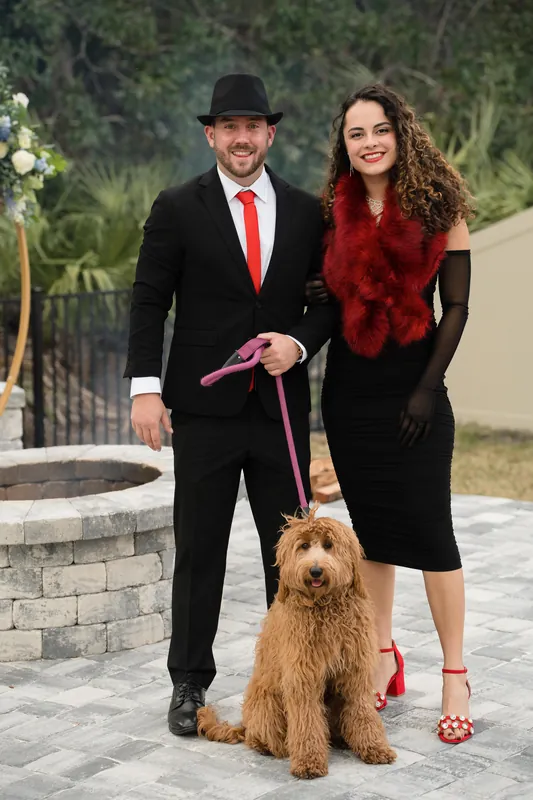 Lea & Marco stand with a golden-brown dog on a paved patio, wearing formal evening wear with festive accents.