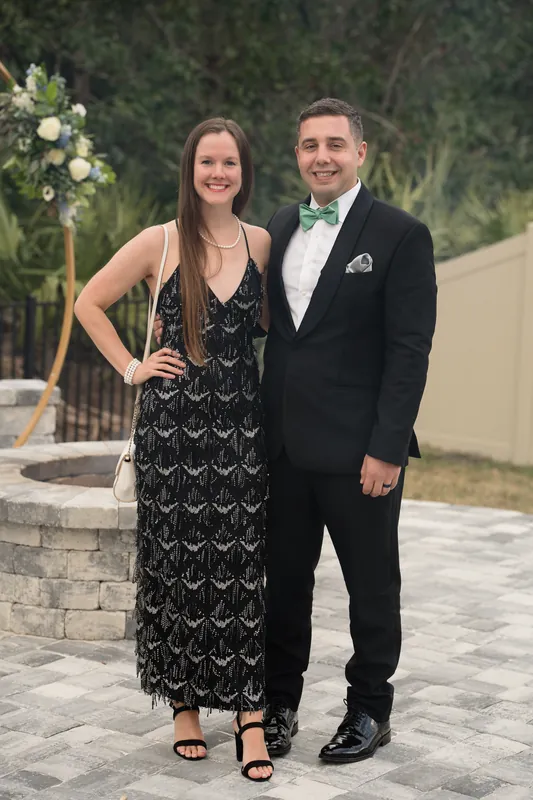 A couple in black formal wear poses together on a stone terrace with flowering plants behind them.