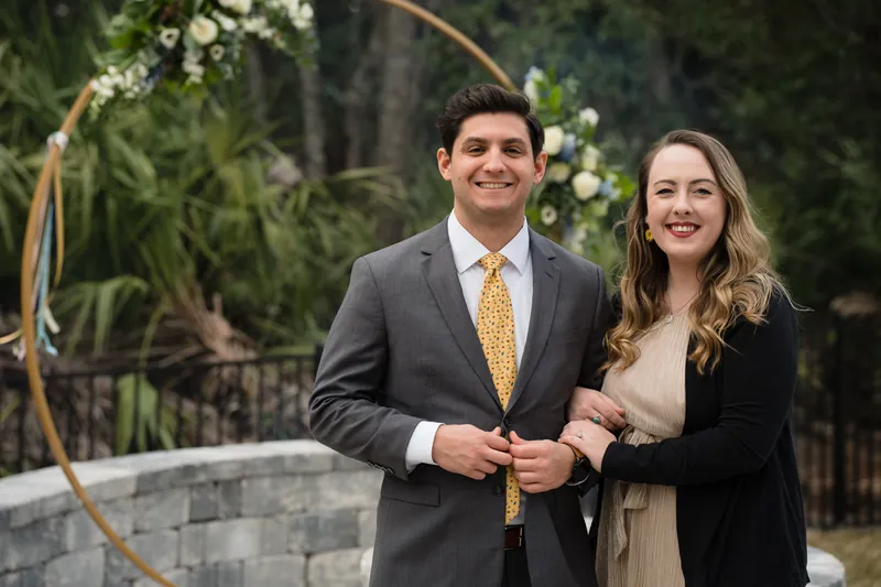 Two guests in gray and cream formal attire stand by a floral arch arrangement on the patio.