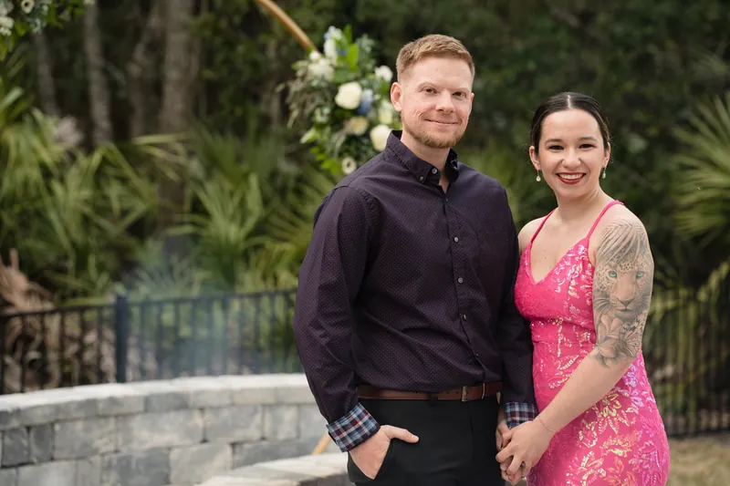 A man in dark formal wear and a woman in a hot-pink patterned dress pose together outdoors near flowering greenery.