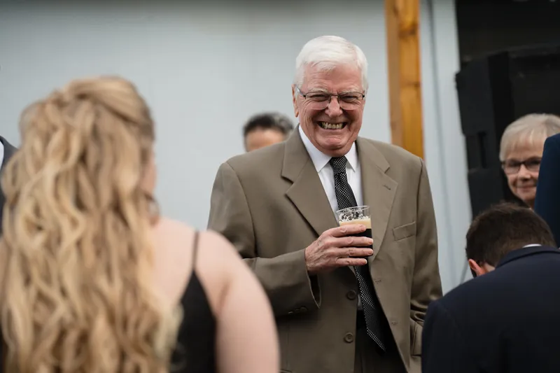 A man in a tan blazer speaks into a microphone while smiling at guests during an indoor reception.