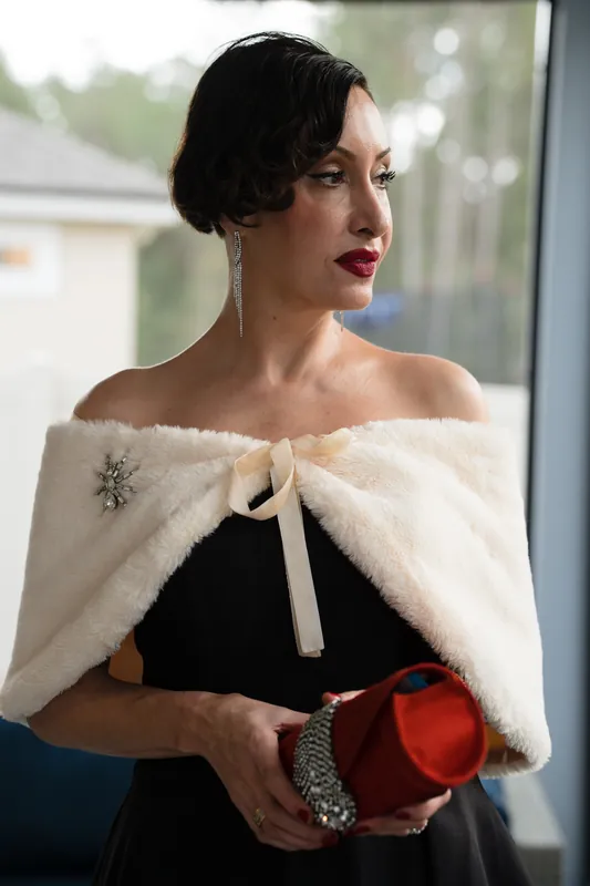 A person in a black dress with a cream fur stole holds a red clutch, gazing to the side near a bright window.