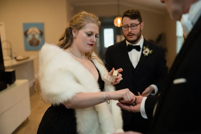 Two people in formal wedding attire—one in a white fur stole over black, the other in a black tuxedo—exchange rings during a ceremony.