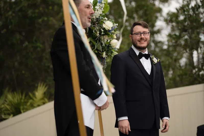 A person in a black coat with white trim stands beside someone in a tuxedo beneath a floral archway outdoors.