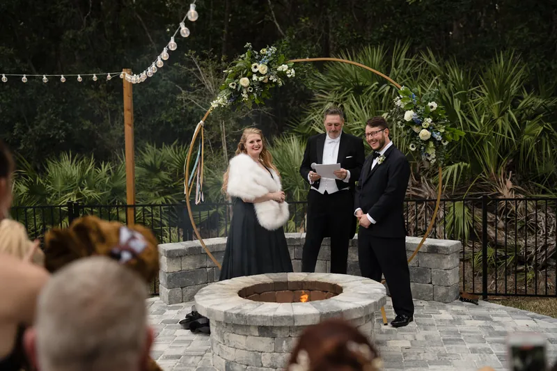 Two people in formal attire stand near a stone fire pit during an outdoor celebration, observed by guests holding drinks.