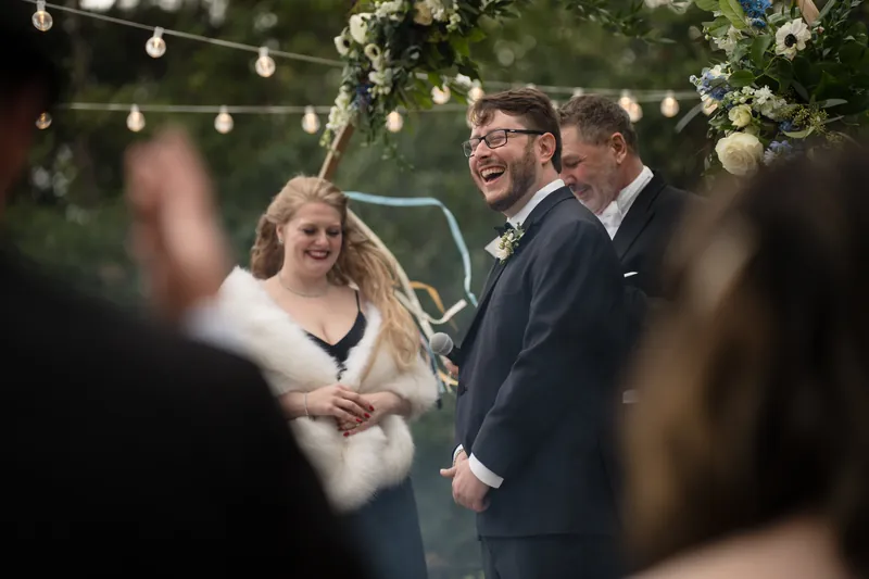 Two people in formal wedding attire smile beneath string lights and floral arrangements, with guests visible in soft focus around them.